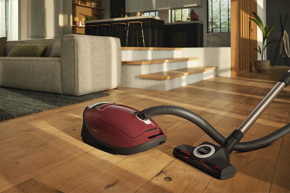A red Miele bagged cylinder vacuum cleaner, hose and floorhead placed on a wooden floor.